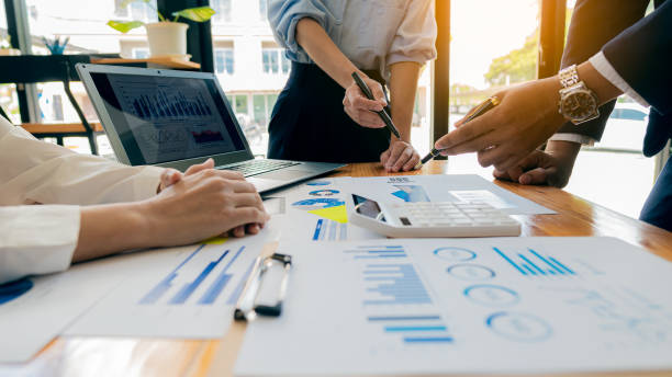 Collaborate at office desks to discuss paperwork. A financial group of business people is meeting to analyze data for a marketing plan on a table with graphs and a laptop with a calculator.