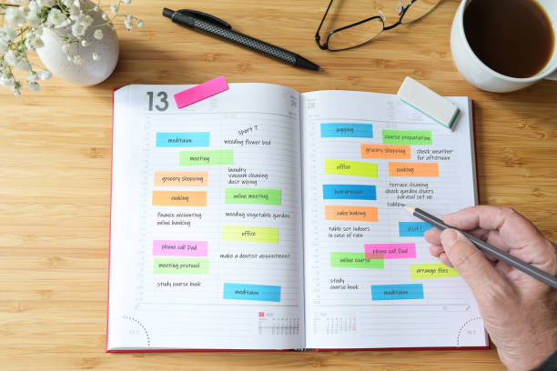 Hand of a woman writing appointments in a diary organizer or schedule calendar with a lot of colorful sticky notes for her time management on a wooden desk with coffee, flowers and glasses, selected focus, narrow depth of field.