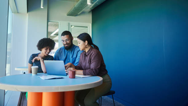 Diverse group of a smiling young businesspeople working together on a laptop during a meeting around a table in an office lounge. Group invite for meetings.